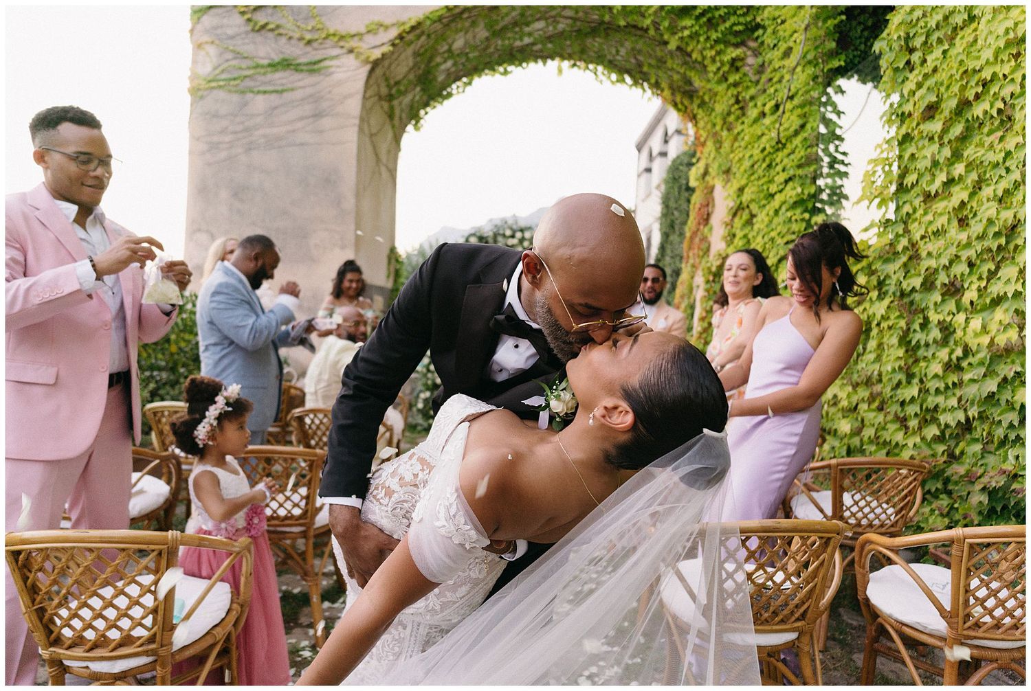A romantic moment captured during an elegant outdoor garden wedding ceremony with guests looking on in pink and formal attire.