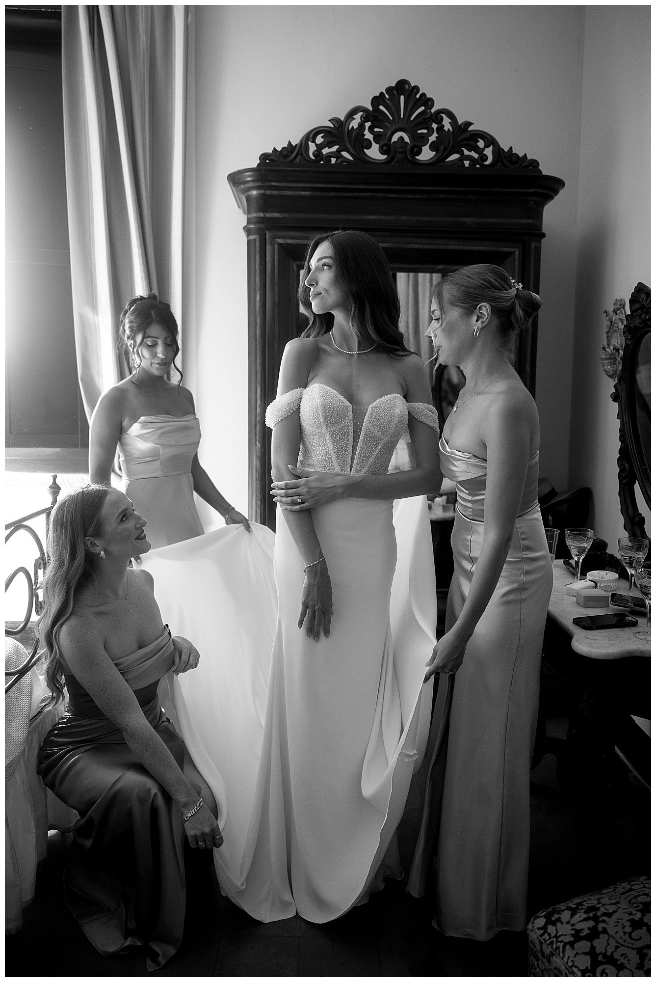 Black and white photo of a bridal party getting ready in an elegant room with an ornate dark wood mirror.