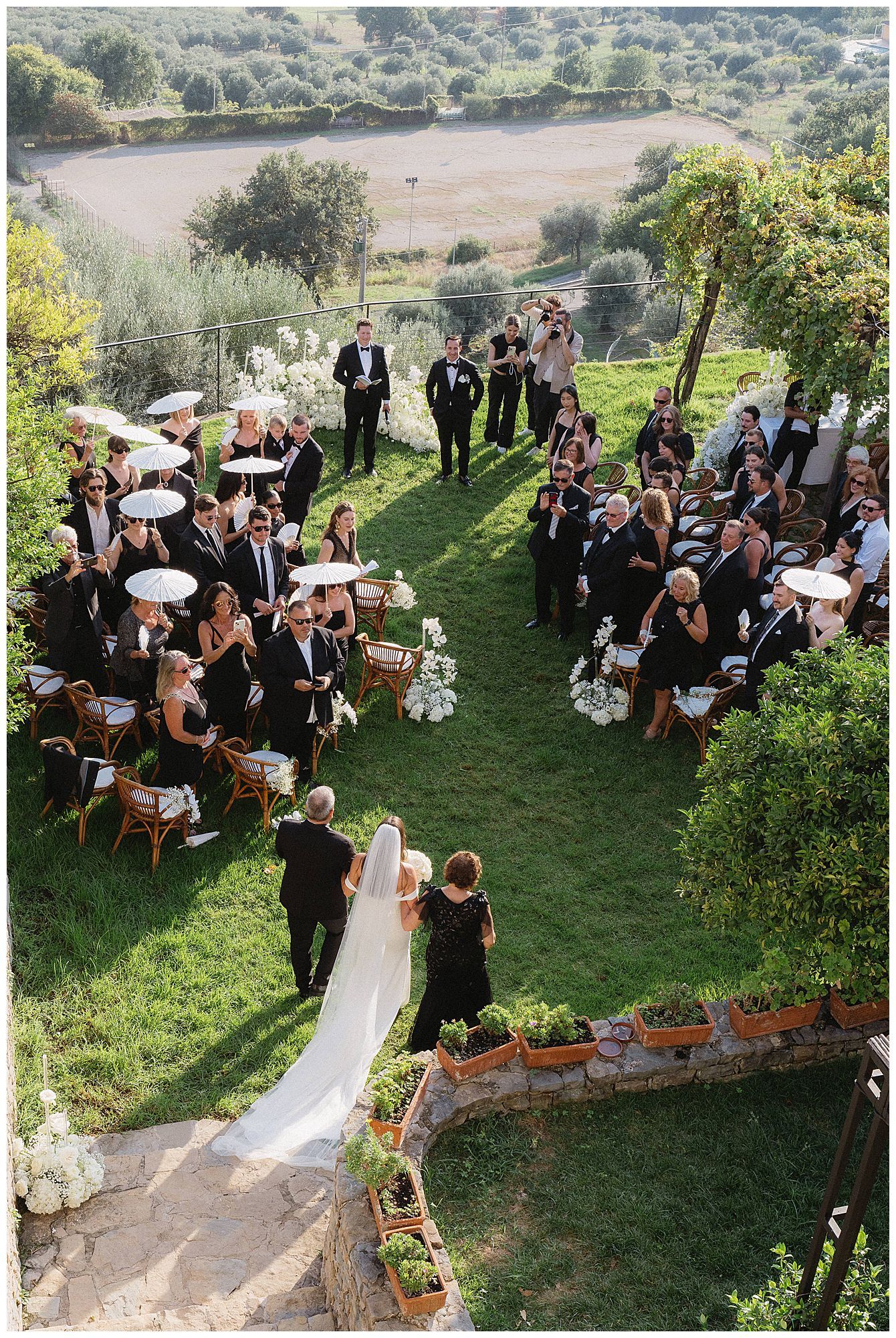 Aerial view of an outdoor wedding ceremony on a terraced garden lawn overlooking a scenic lake in the countryside.