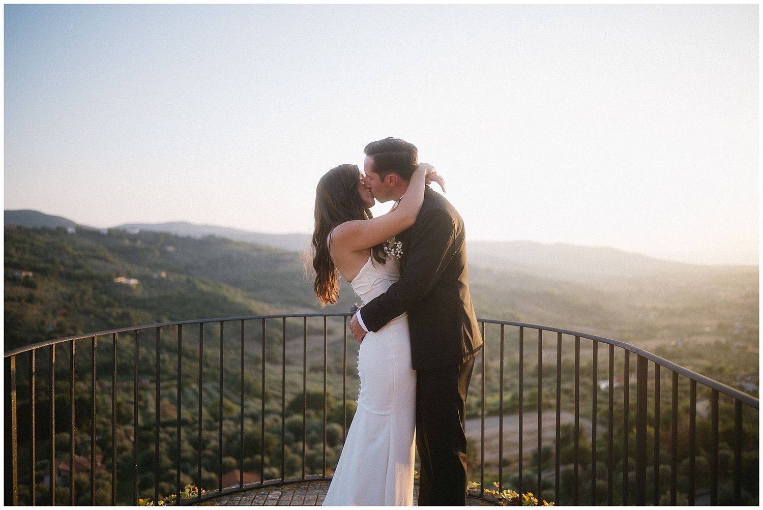 Couple in wedding attire embraces against backdrop of rolling hills and mountain vista at sunset.