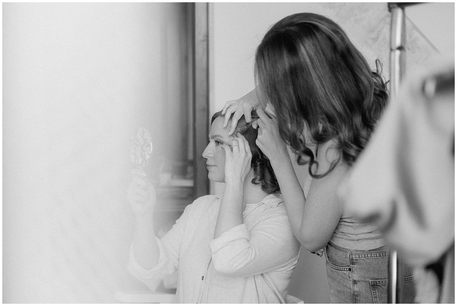 A candid black and white moment of someone having their hair styled in a bridal preparation setting.