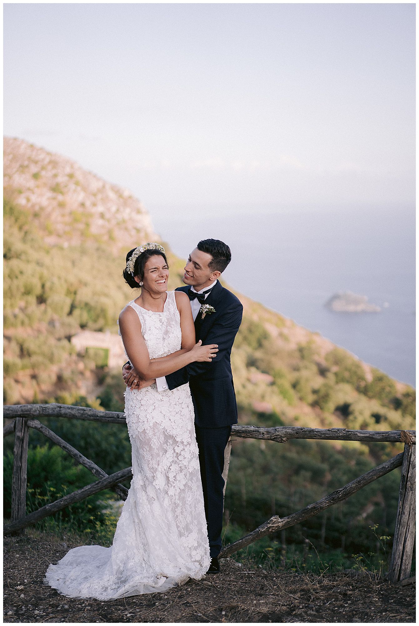A couple shares a romantic moment in lace wedding dress and black tuxedo on a coastal hilltop with ocean views at sunset.