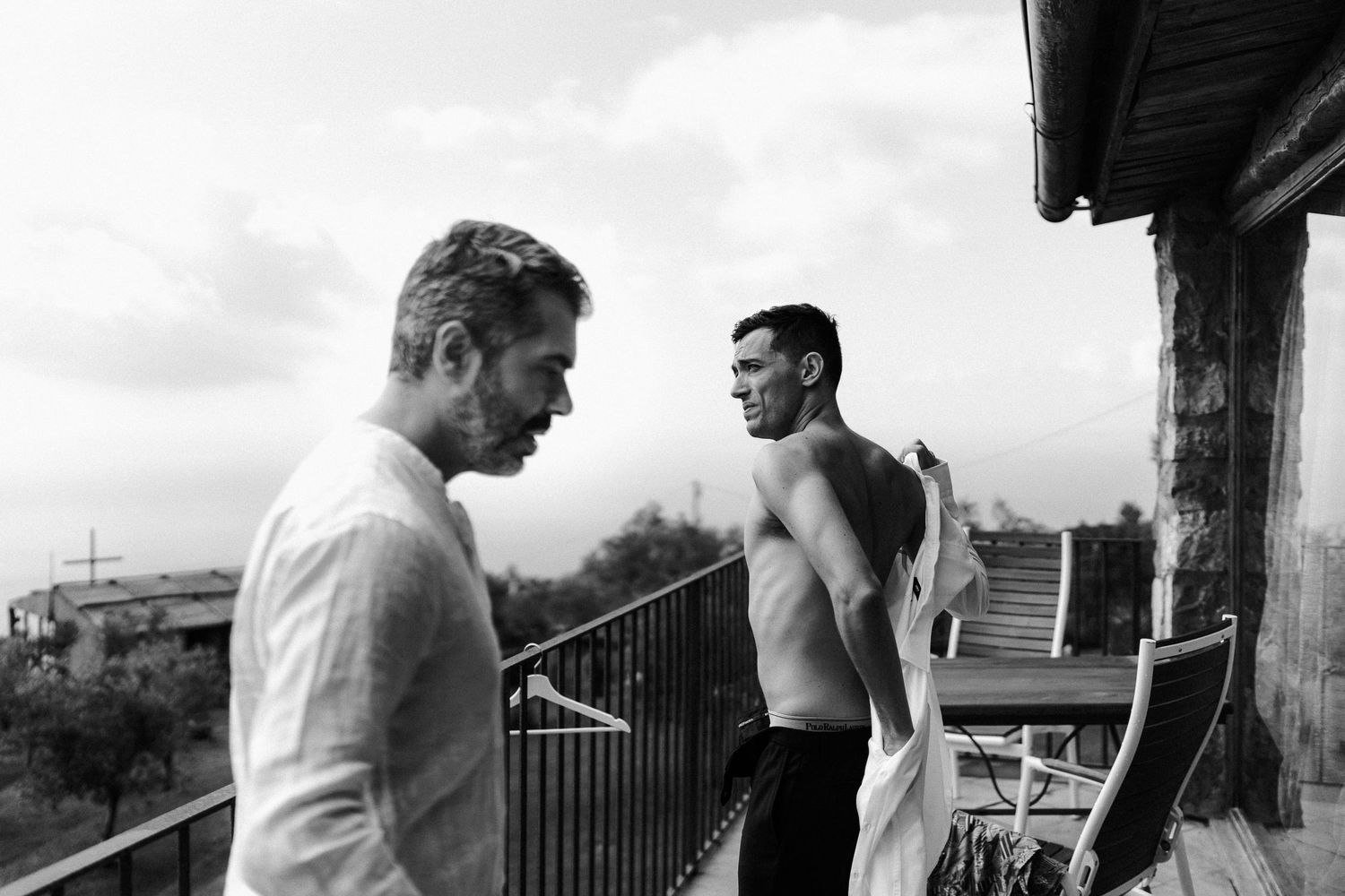 Two figures in black and white stand on a balcony, one wearing a white shirt while the other is shirtless.