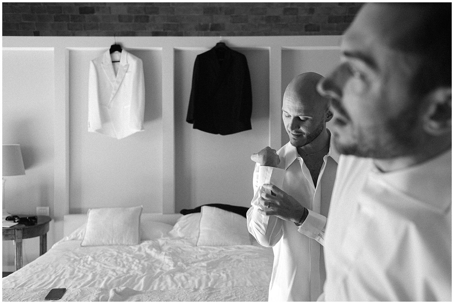 A black and white photograph showing formal attire hanging in a bedroom with light streaming through a window.