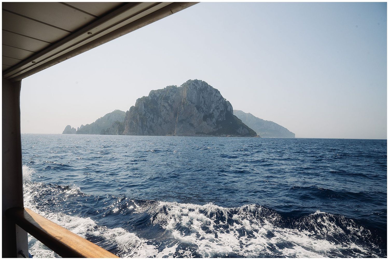 Rocky island mountain rises from blue Mediterranean waters viewed from moving boat creating white wake patterns.