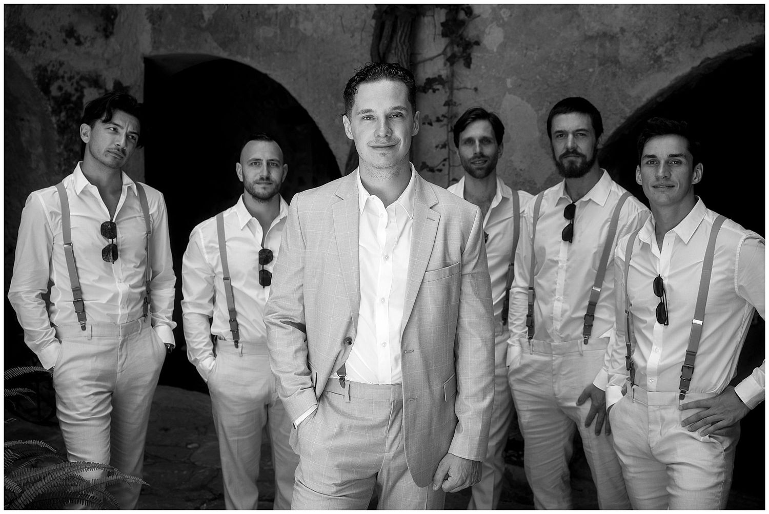 Groomsmen in matching white formal attire pose together in black and white photo against stone wall backdrop.