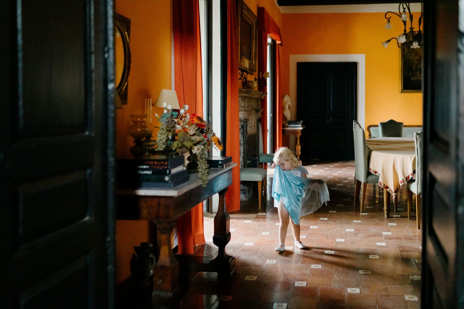 A small child in a blue dress explores a sunlit interior room with orange walls and antique furniture.