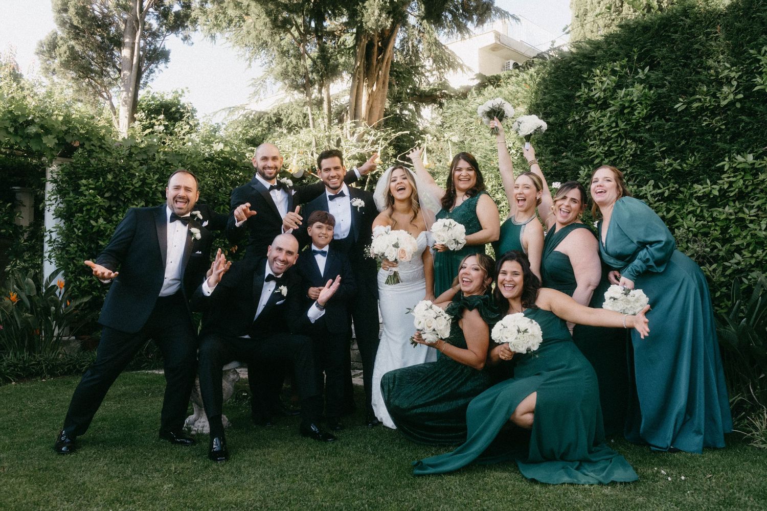 Wedding party poses for fun group photo in garden wearing black and emerald green attire.