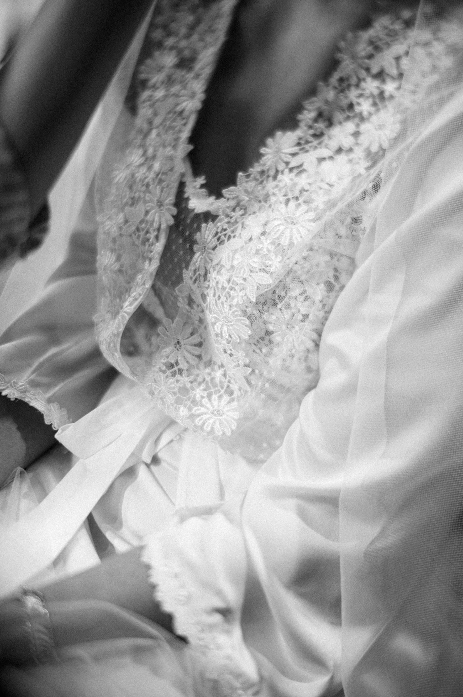 Close-up black and white detail of delicate white lace and fabric from a wedding dress showing intricate patterns and folds.