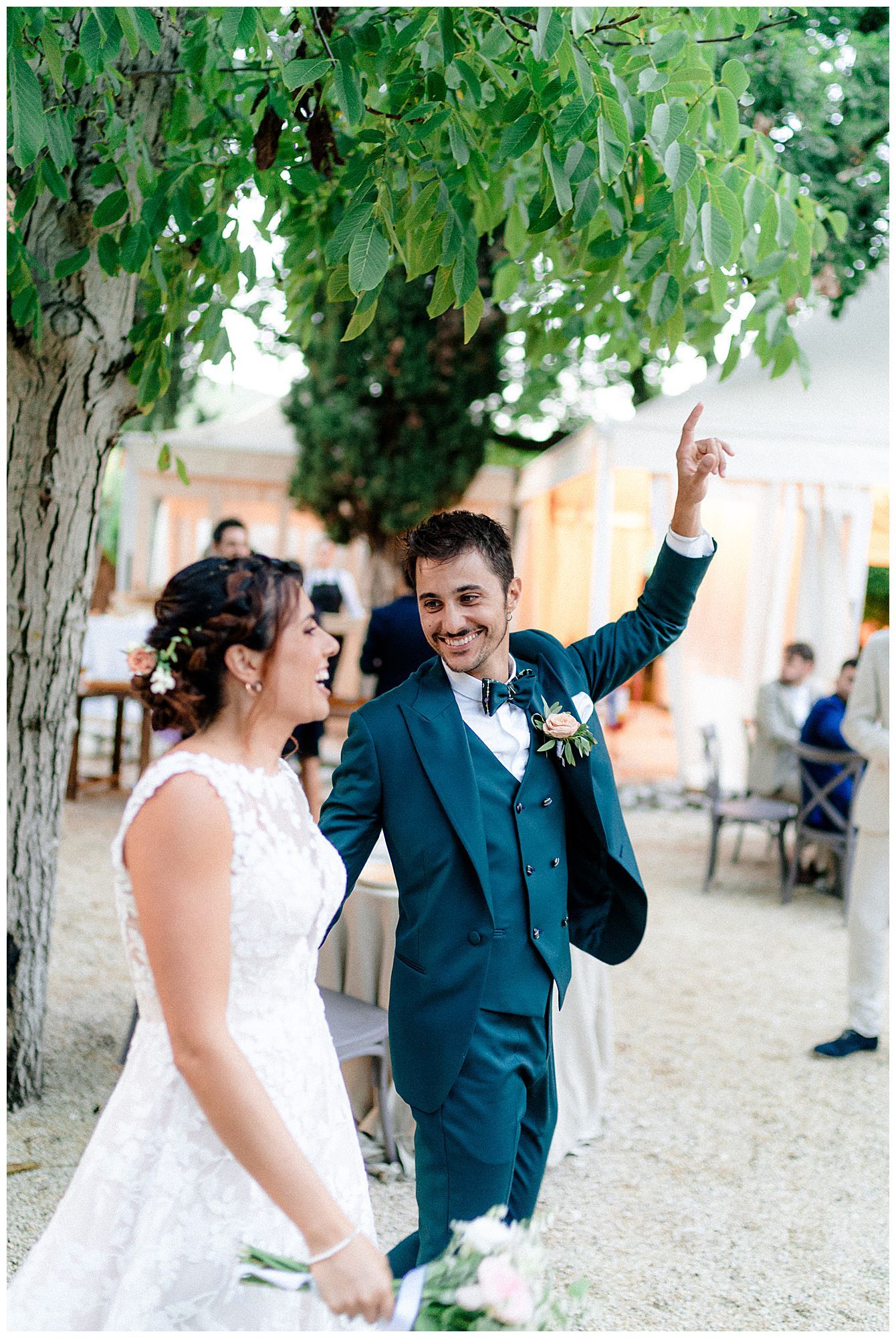 Joyous couple celebrates during outdoor wedding reception under green tree canopy with rustic venue setting in background.