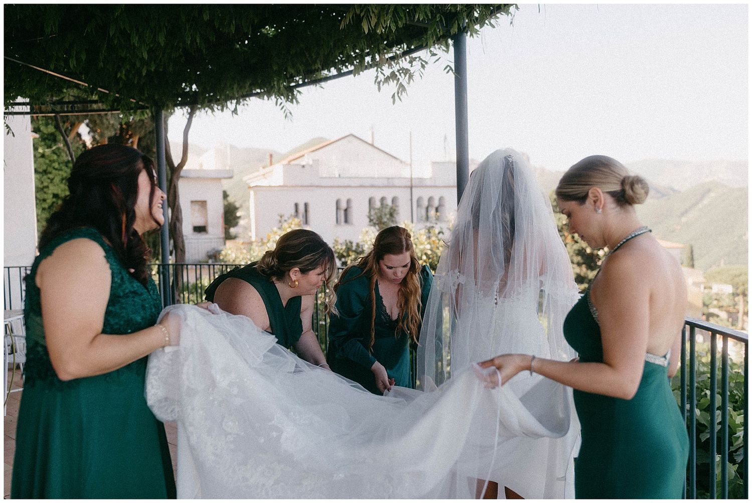 A group in emerald green dresses help adjust a delicate white wedding veil on an outdoor balcony overlooking distant hills.