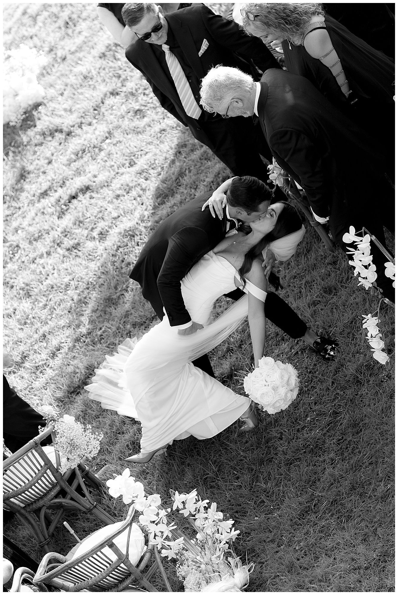 Aerial black and white photo of newlyweds dipping on grass surrounded by wedding guests and flower arrangements.