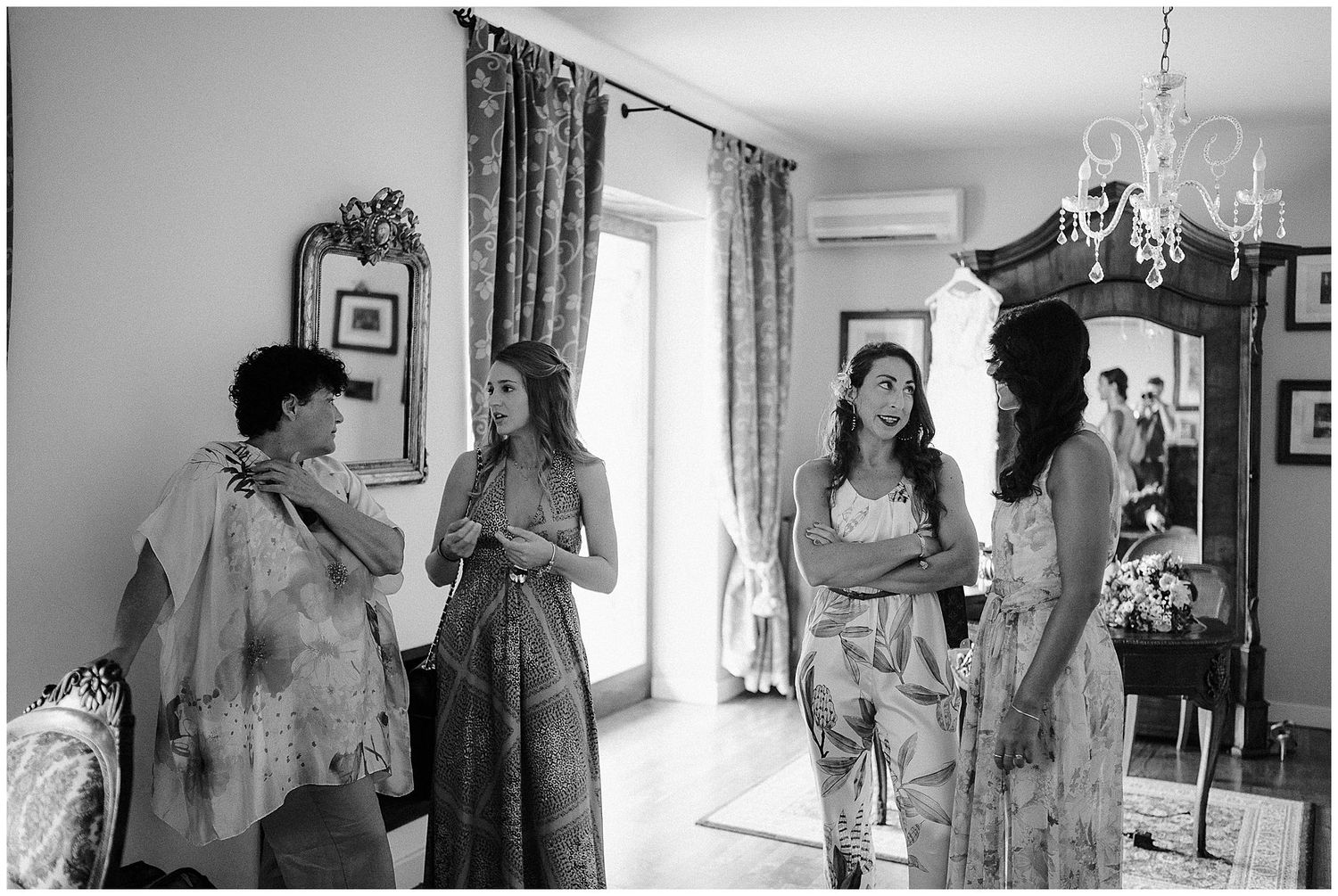 A candid black and white photograph showing a group of people in conversation inside an elegant room with curtains and decor.