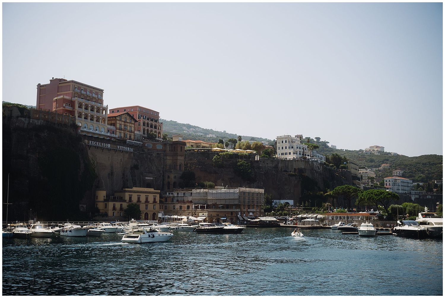 Small boats line the harbor against a hillside with Mediterranean buildings and houses overlooking the water in Sorrento, Italy.