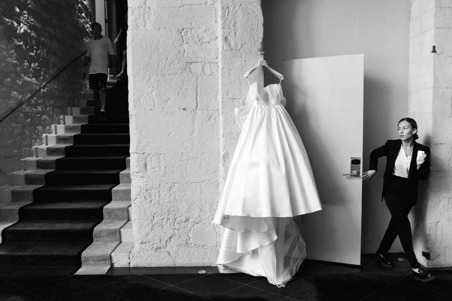 A wedding dress hangs elegantly on a white wall while a figure stands in the shadows near a staircase.