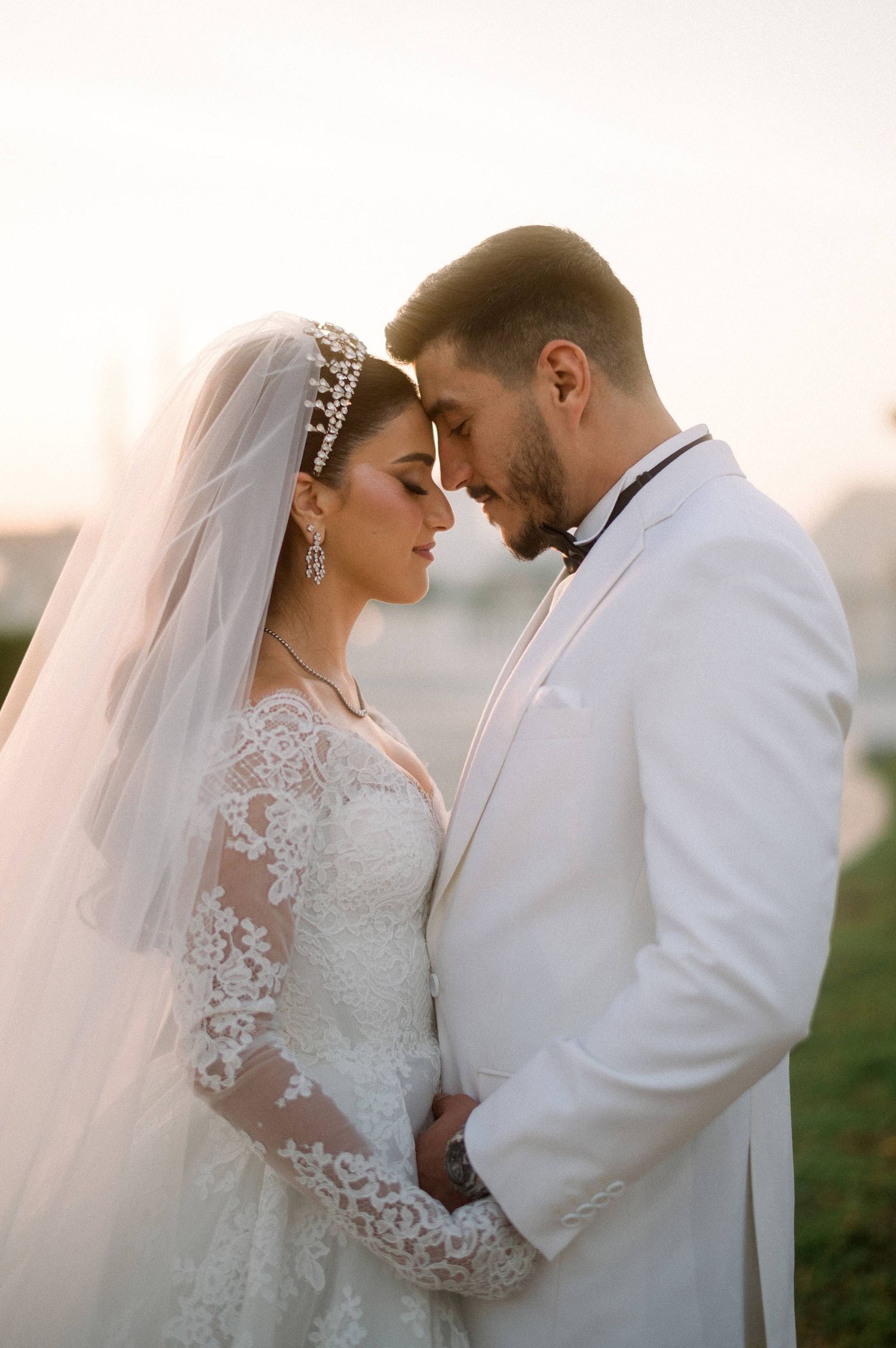 Romantic wedding portrait of a couple embracing outdoors at sunset in elegant white attire with lace details and flowing veil.