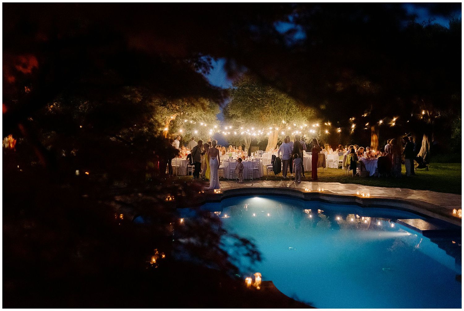 Elegant outdoor evening wedding reception with guests gathered around an illuminated pool under string lights.