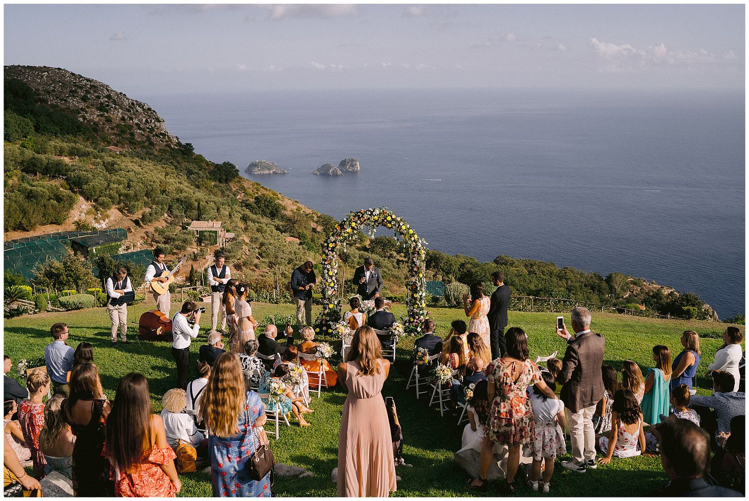 Outdoor wedding ceremony overlooking dramatic coastal cliffs and ocean views with guests gathered on green hillside.