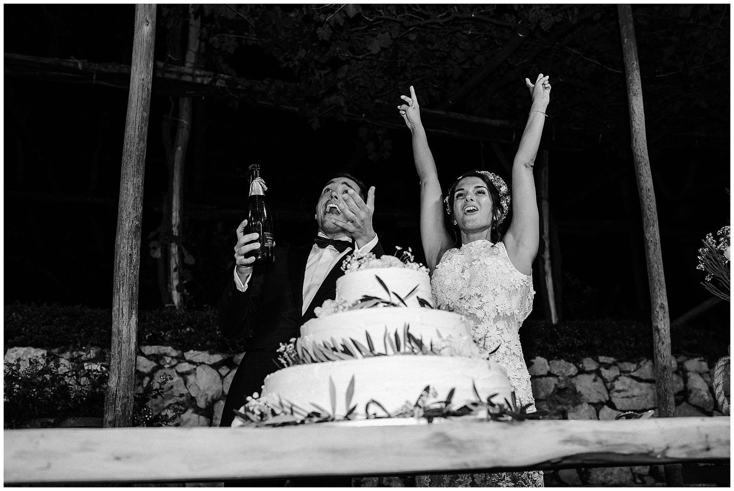 Two figures in wedding attire sit and raise their hands joyfully in a black and white candid moment.