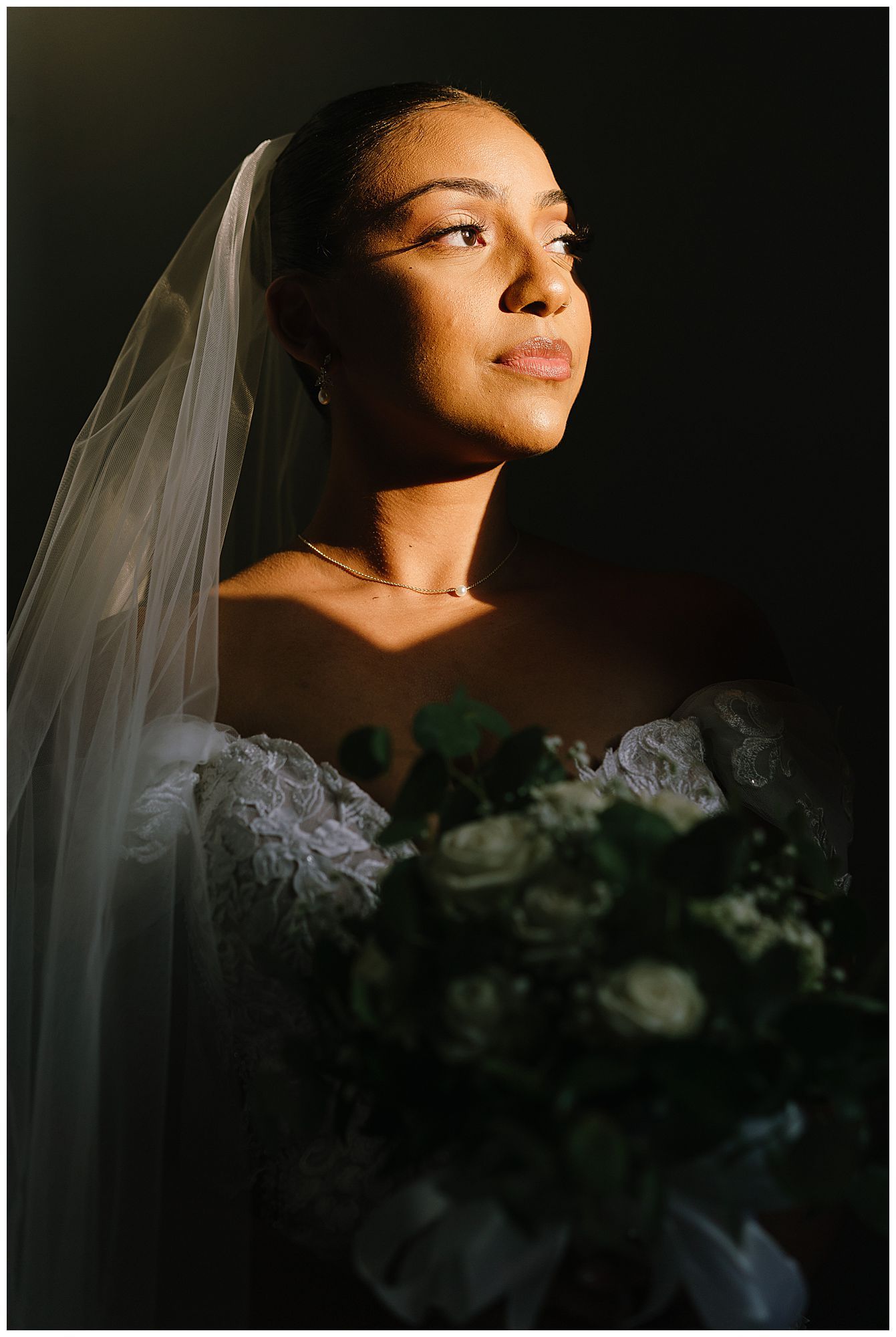A bride in a white veil poses for a dramatic portrait with moody lighting while holding a bouquet in shadow.