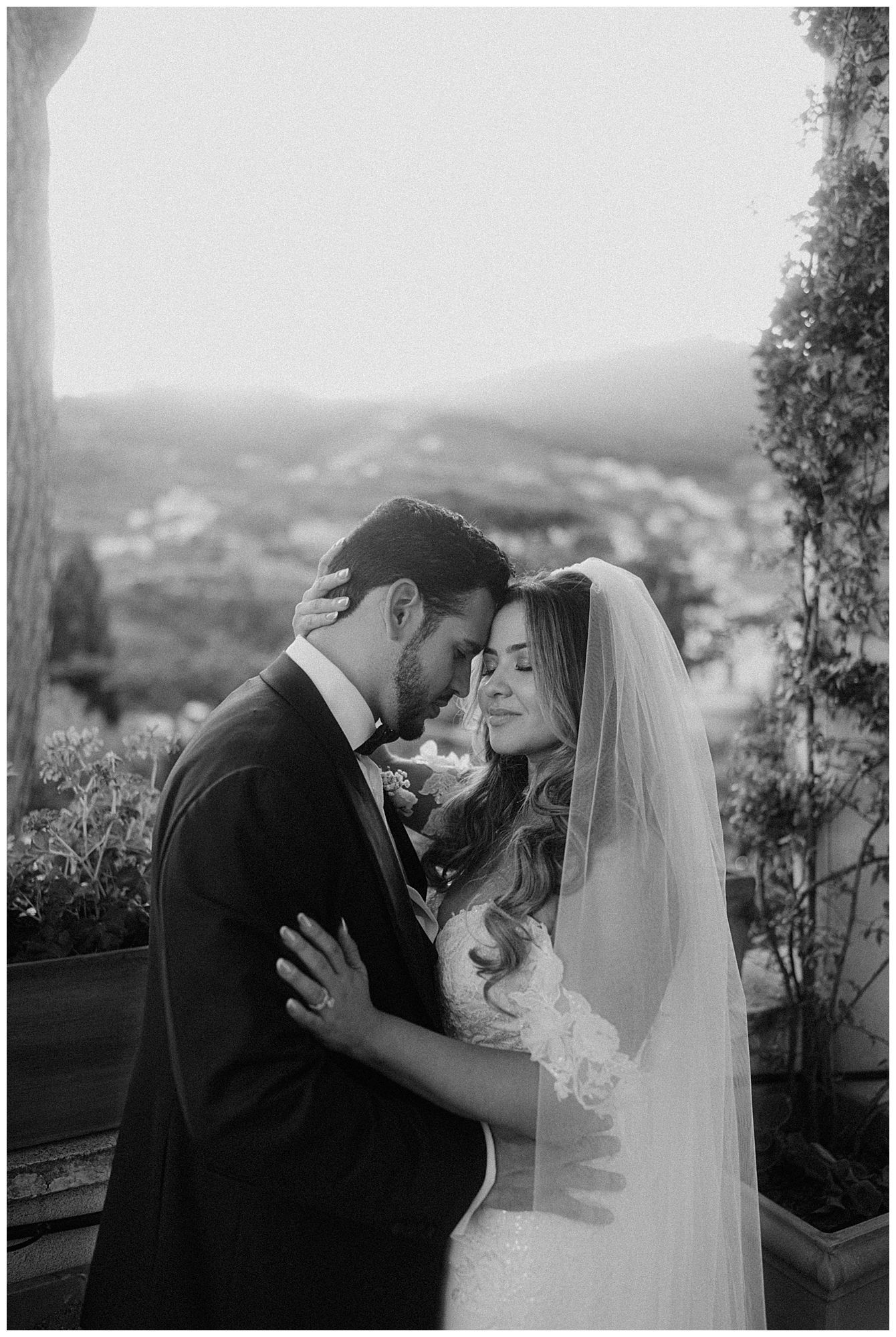 A romantic black and white wedding photo of a couple sharing an intimate moment on a balcony overlooking a scenic view.