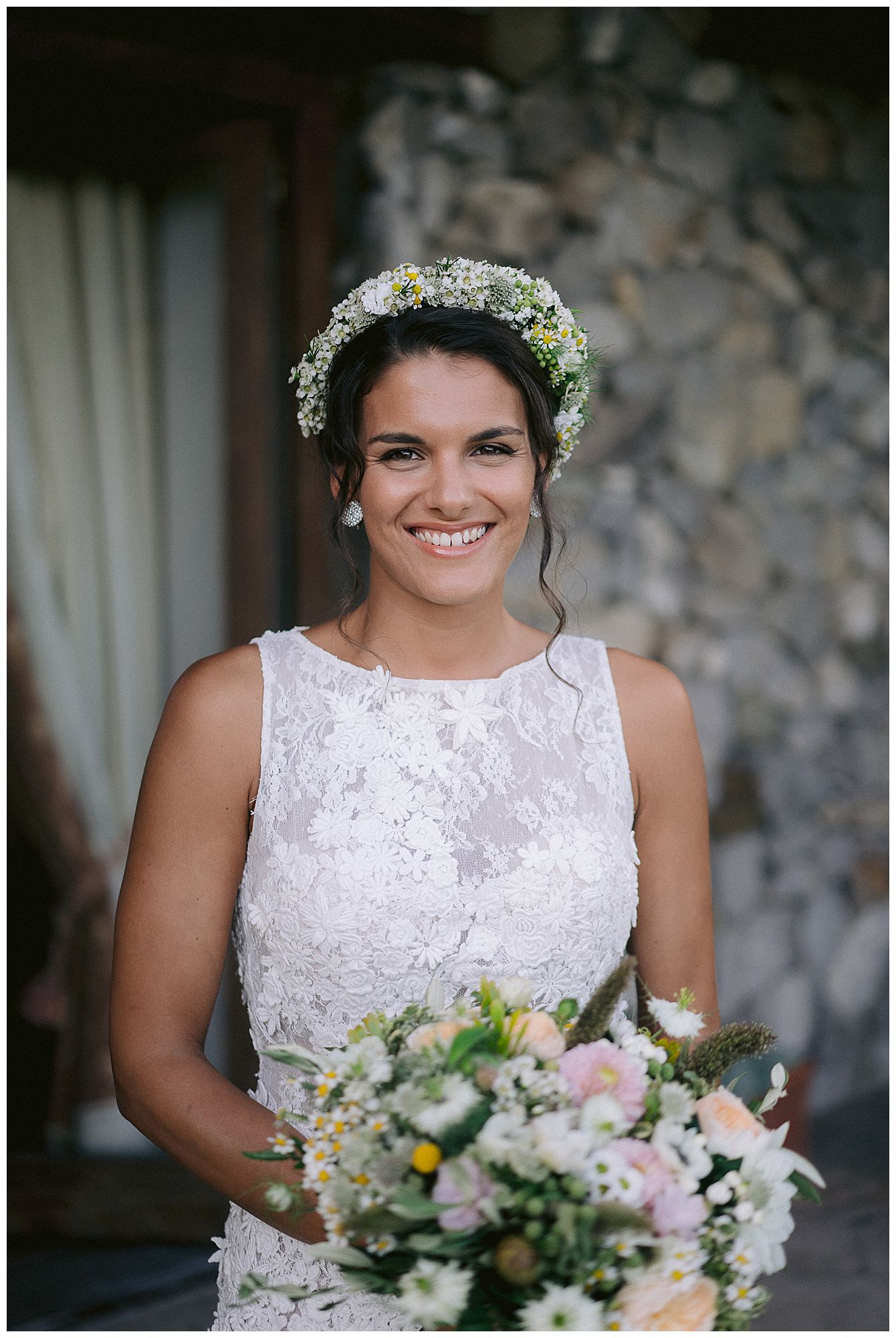 A bride in a white lace dress and floral crown holds a pastel bouquet while posing against a stone wall background.