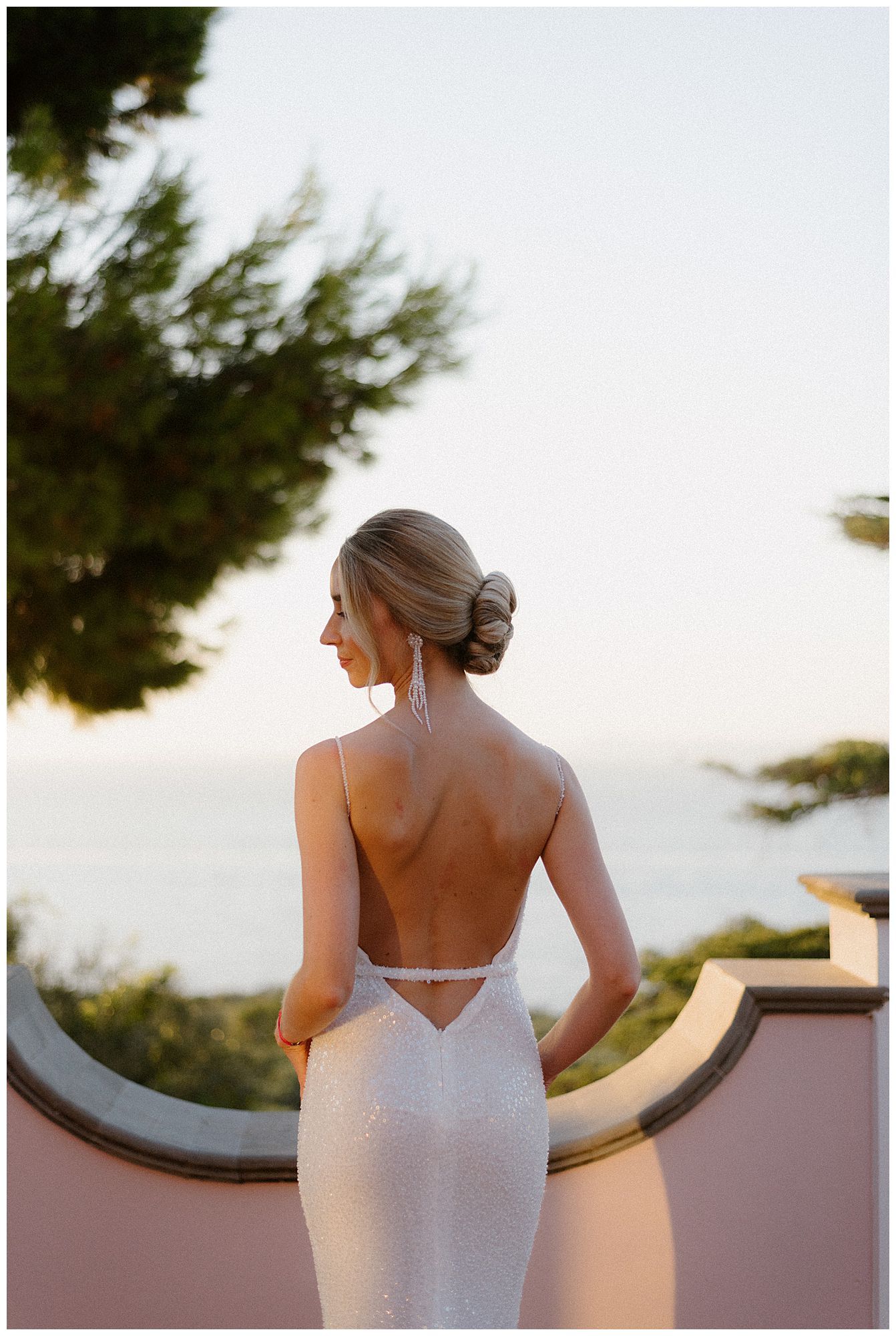 An elegant white backless wedding dress with a fitted silhouette captured at sunset against a curved architectural detail.