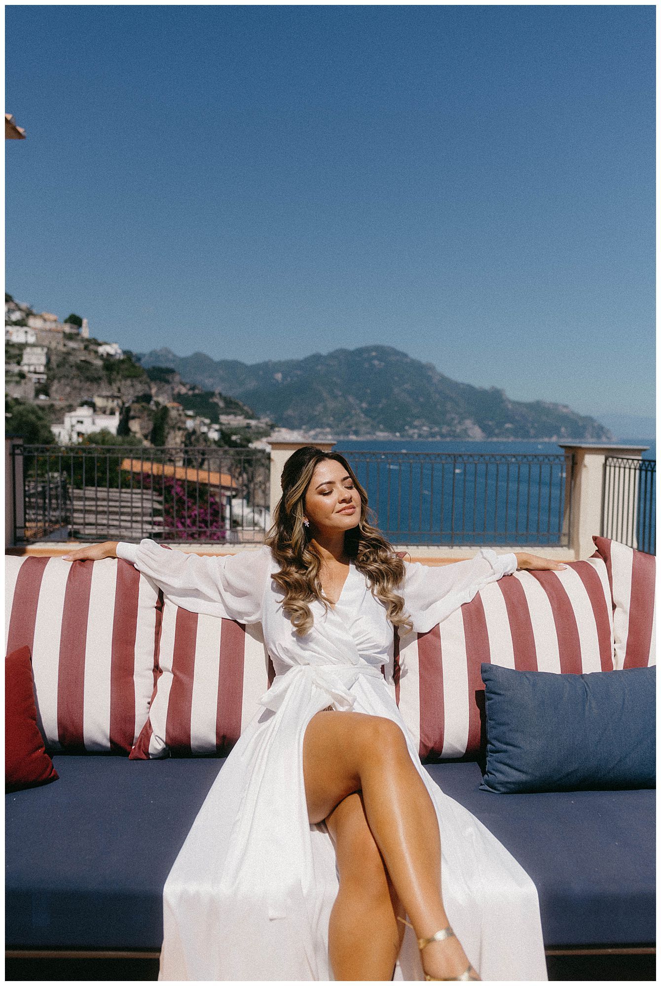 Woman in white dress relaxing on striped cushions with Amalfi Coast Mediterranean view from luxury terrace in Italy.