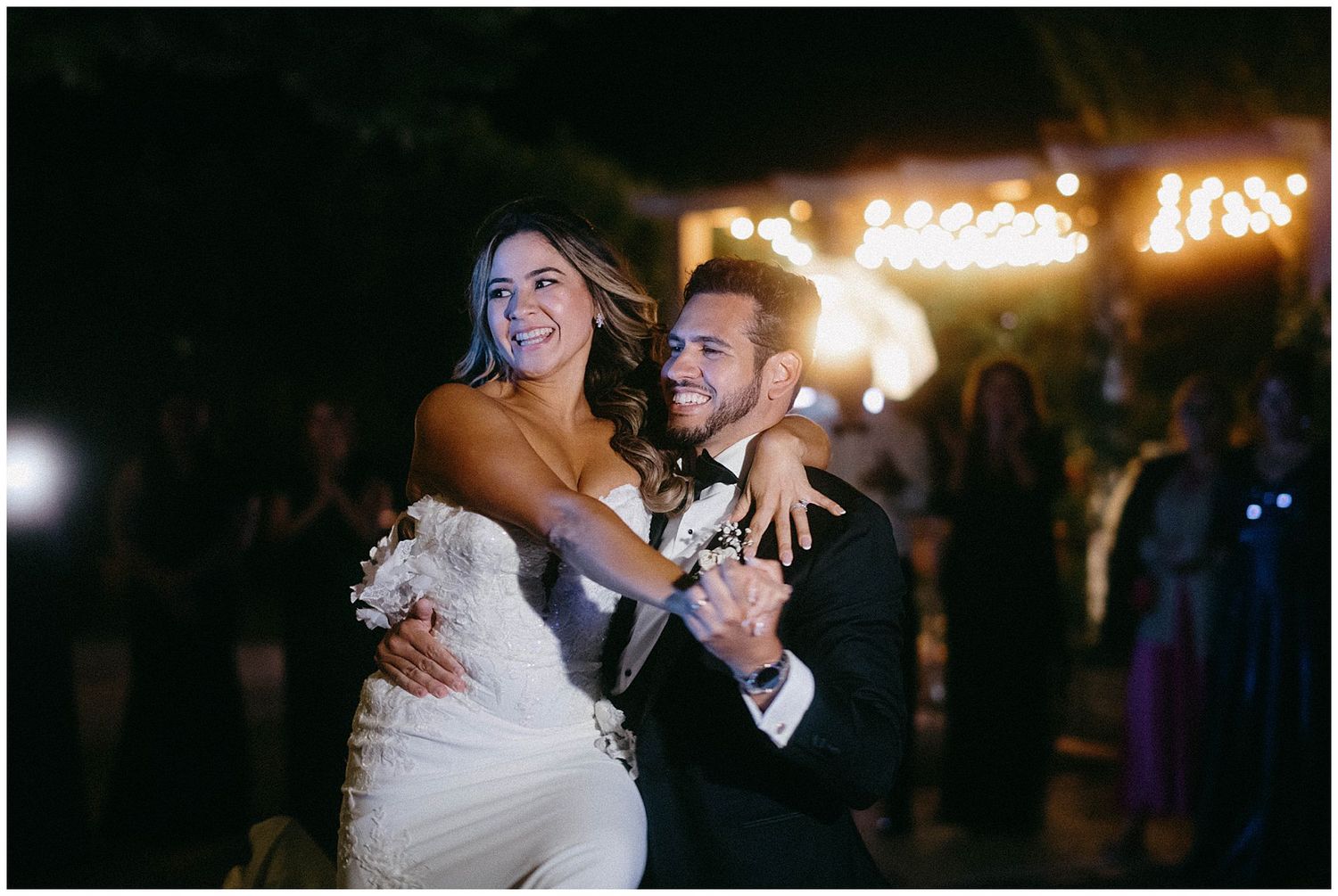A joyful couple dances at their wedding reception while guests look on in the background with twinkling lights overhead.