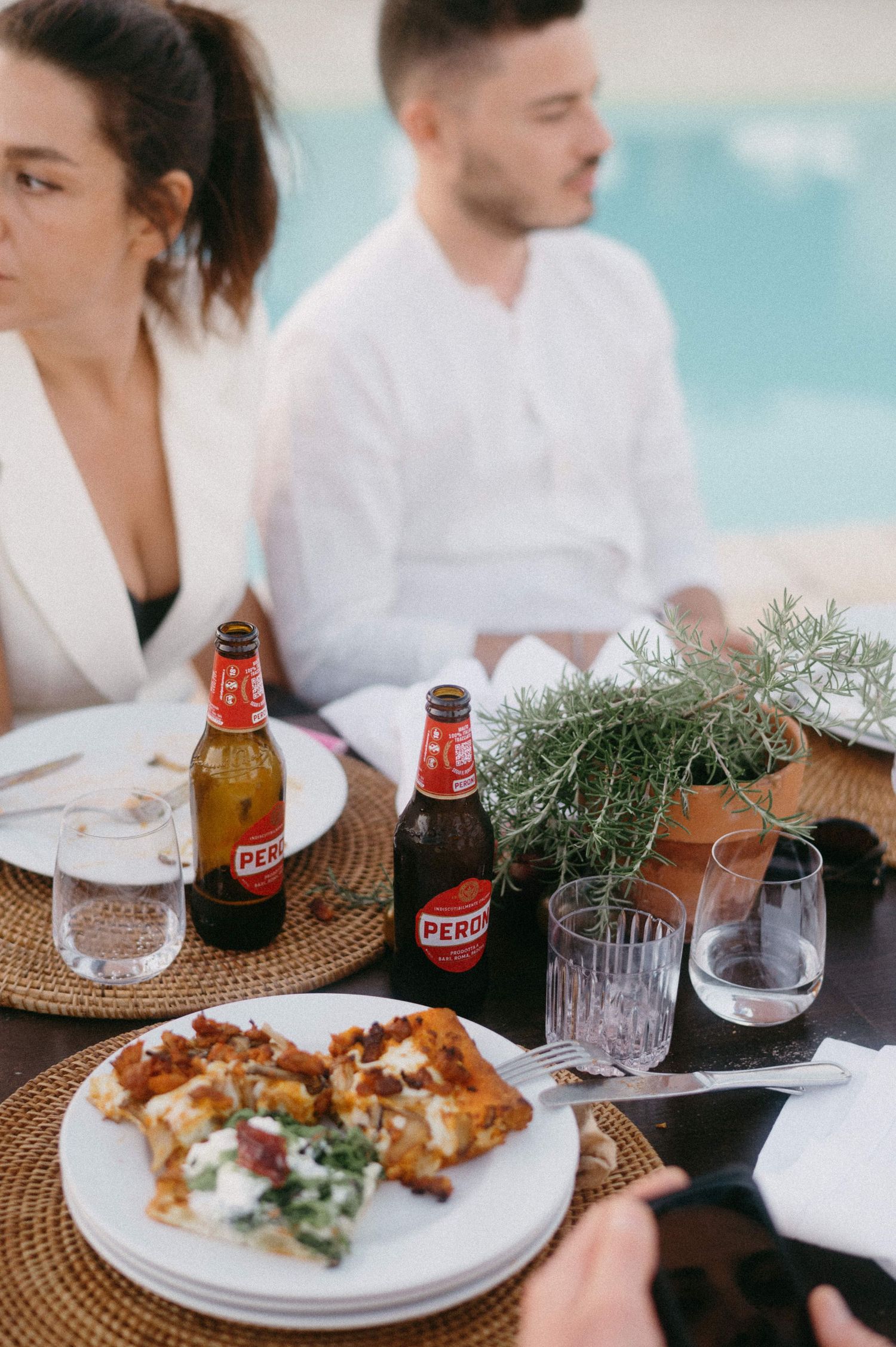 A couple enjoying an outdoor meal with red beer bottles and food on woven placemats by a turquoise backdrop.