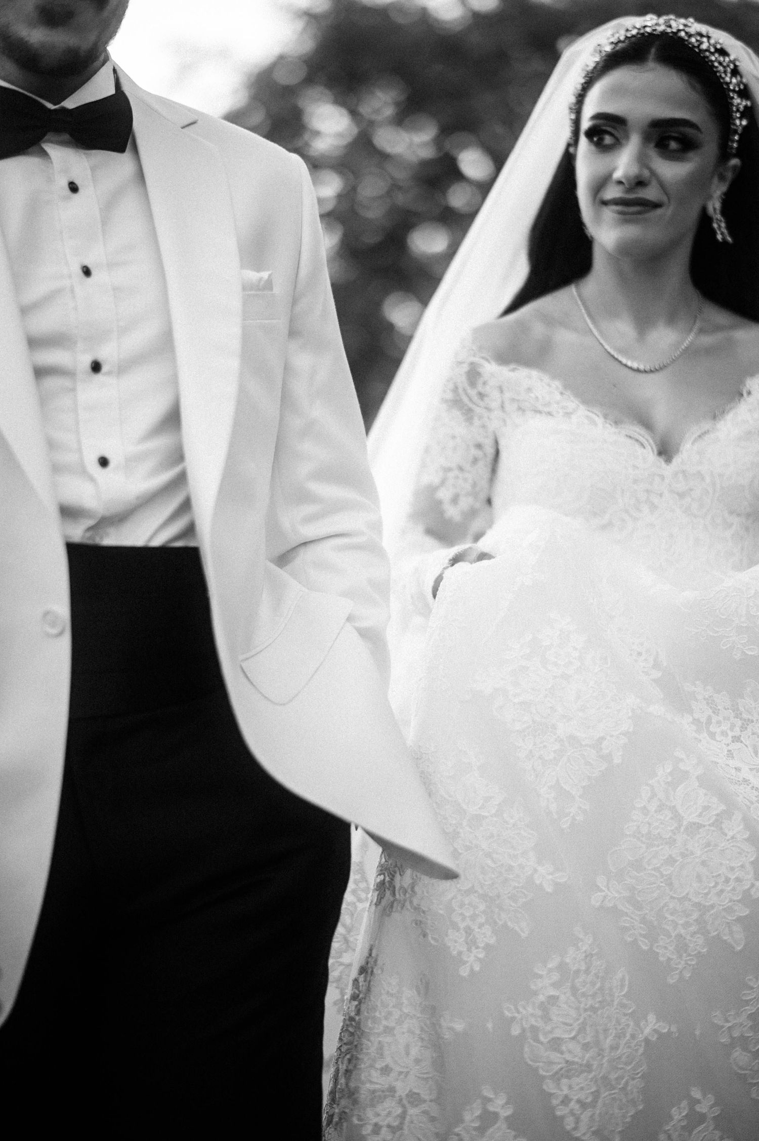 Black and white wedding photo with bride in lace gown and veil alongside groom in white tuxedo jacket.
