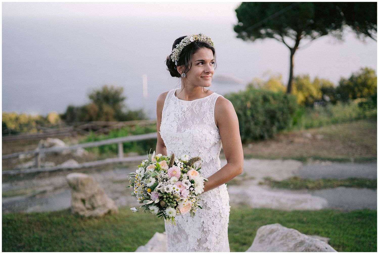 A person in a white lace wedding dress holds an elegant bouquet of mixed flowers on a grassy hilltop with trees in background.