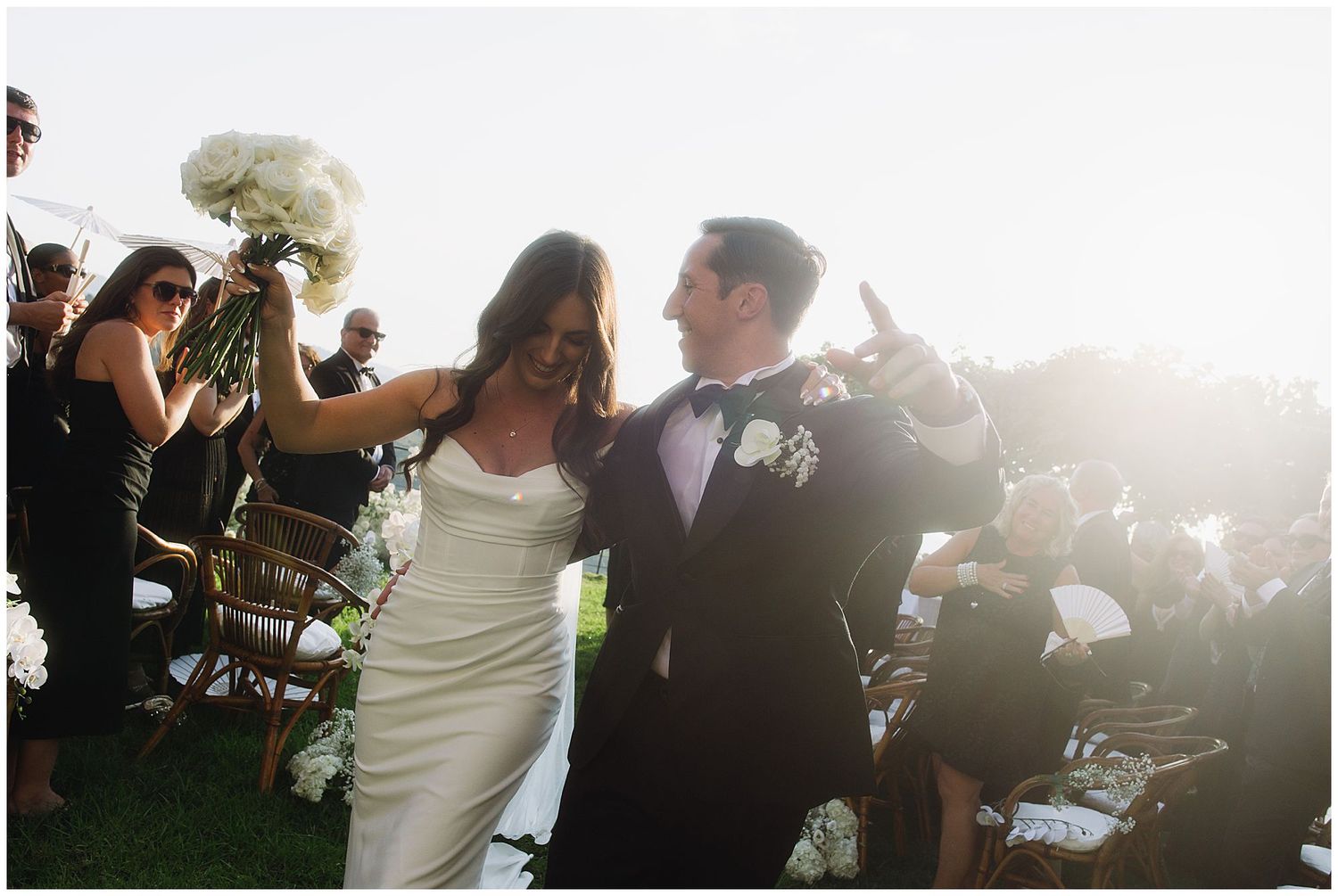 A bride and groom walk together through their outdoor wedding celebration surrounded by guests and sunlit ambiance.