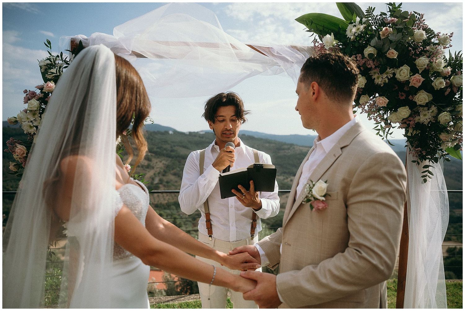 Outdoor wedding ceremony under floral arch with couple holding hands as officiant reads from book.