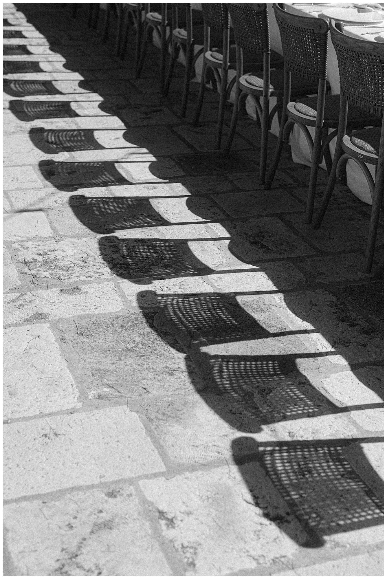 A black and white photograph of chair shadows casting geometric patterns on a stone floor.