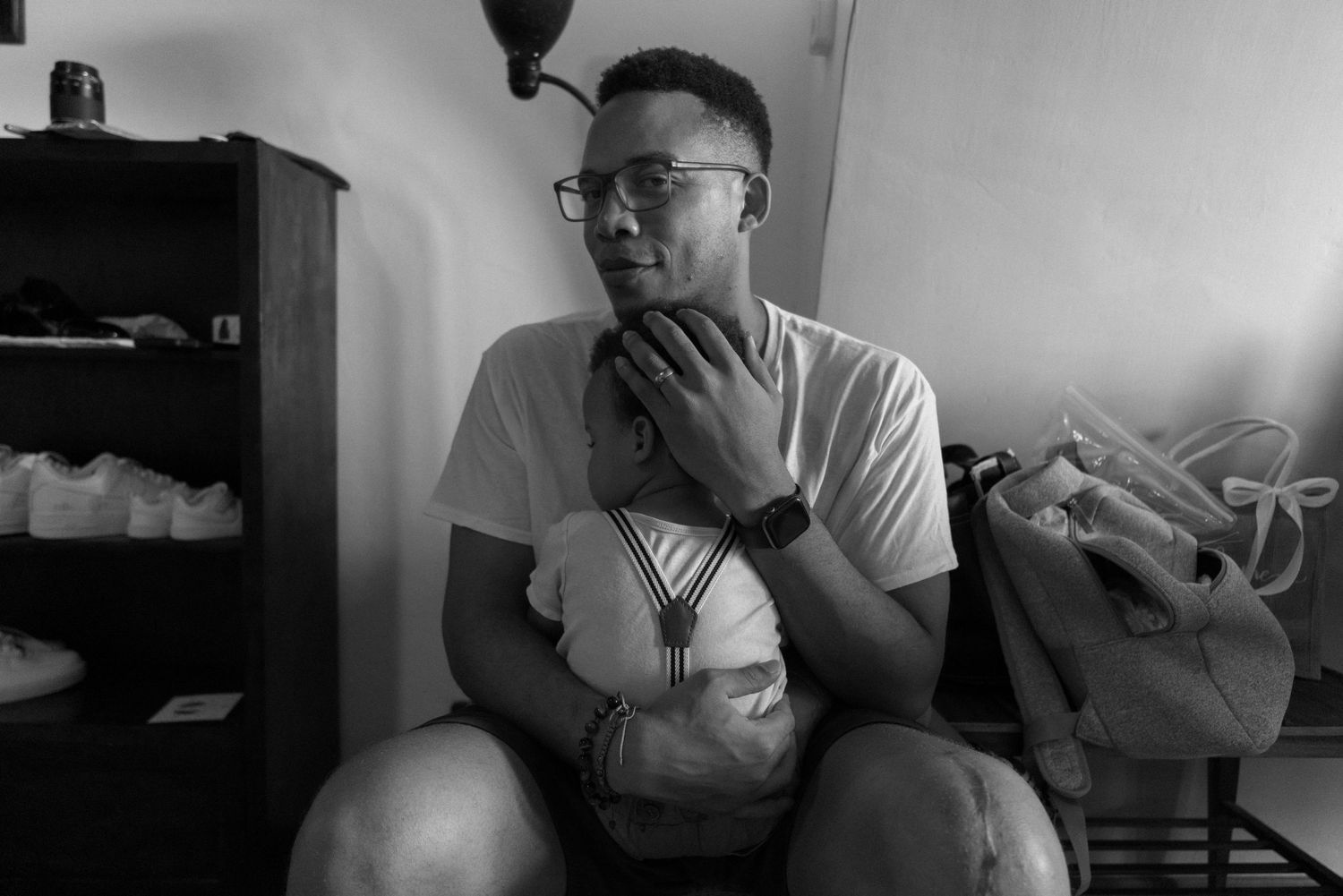 Black and white candid photo showing a person in glasses and a white t-shirt sitting thoughtfully in a room.