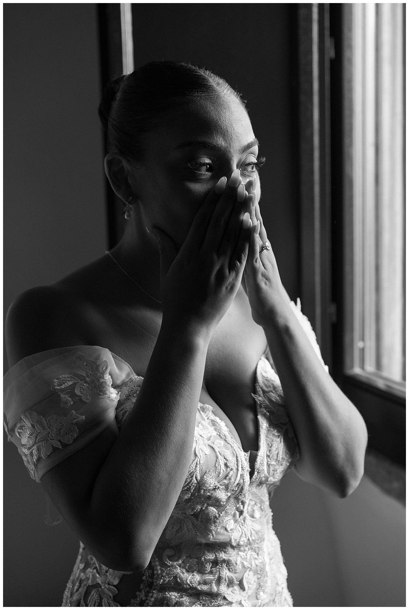 Black and white portrait of an emotional person in a elegant lace wedding dress covering their mouth by a window.