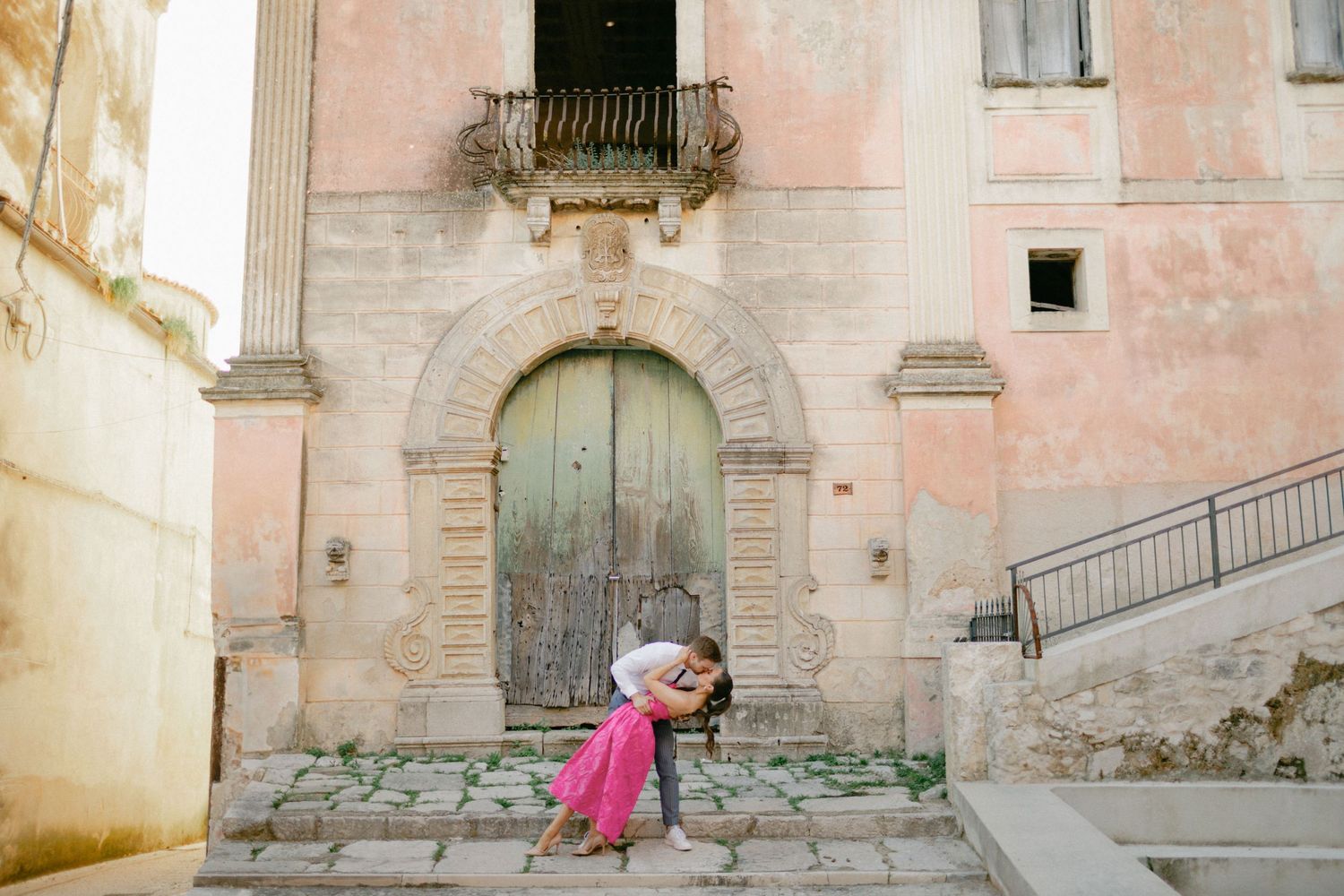 A romantic moment between a couple in front of a pink Mediterranean building with an arched doorway.