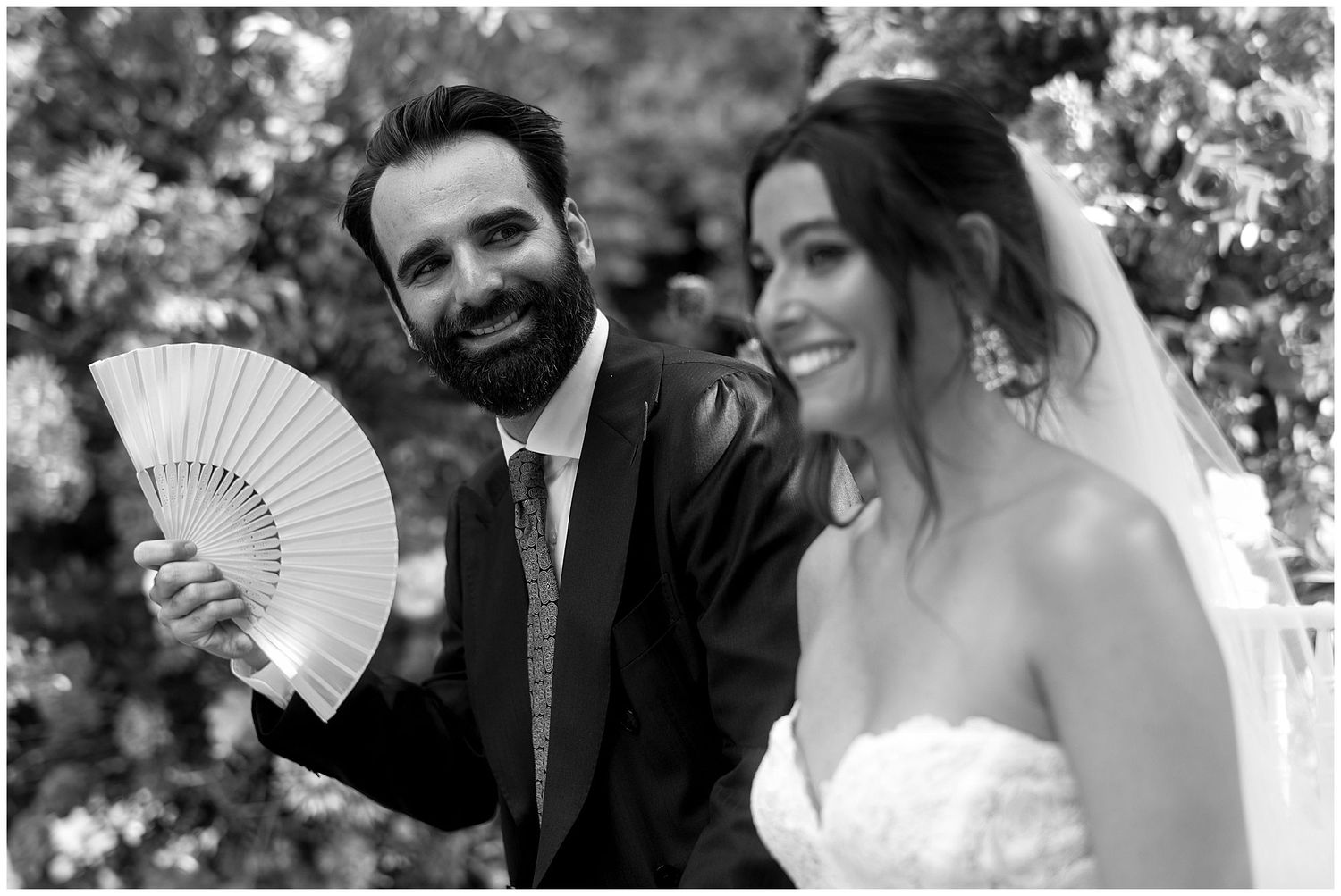 A couple in wedding attire shares a joyful moment outdoors with a decorative white hand fan.