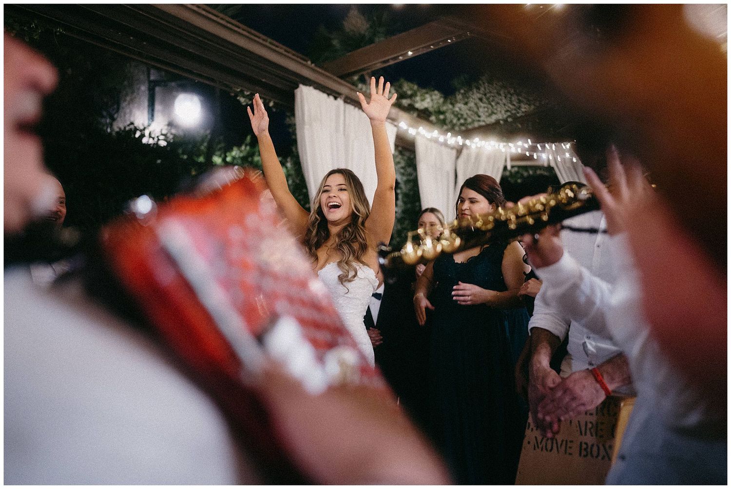 Energetic crowd dances with raised arms at a lively nighttime celebration under string lights and decorations.