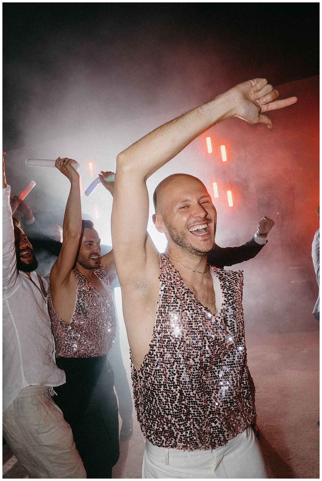 Energetic partygoers dancing and celebrating in sparkly attire under atmospheric red lighting and smoke effects.