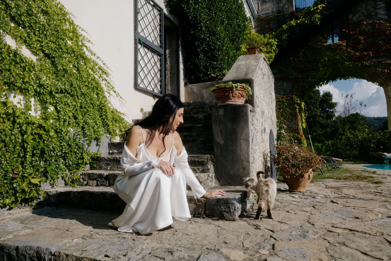 A woman in a white dress sits on stone steps against an ivy-covered wall.