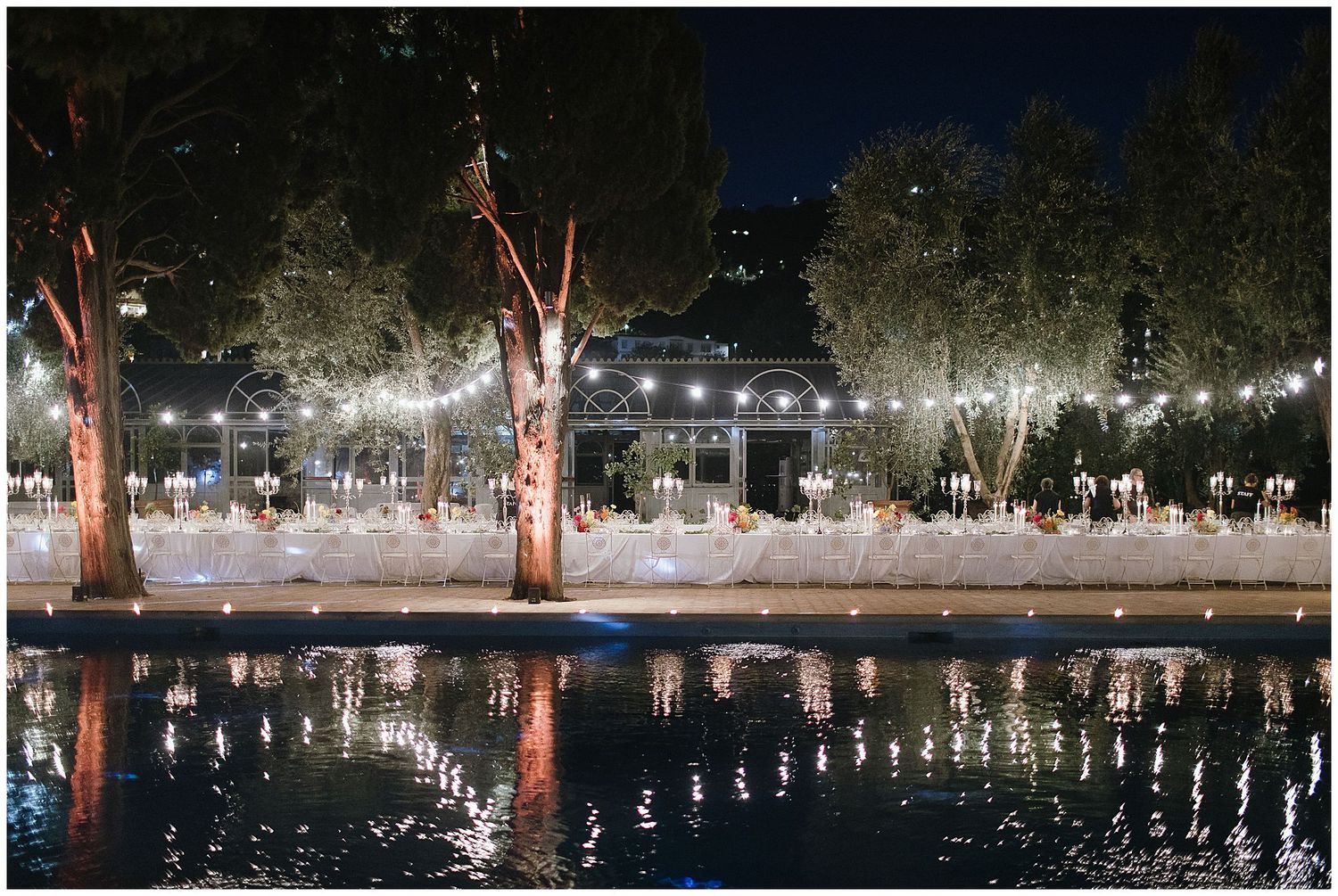 String lights illuminate a nighttime outdoor venue with trees reflecting beautifully in a calm pool of water.