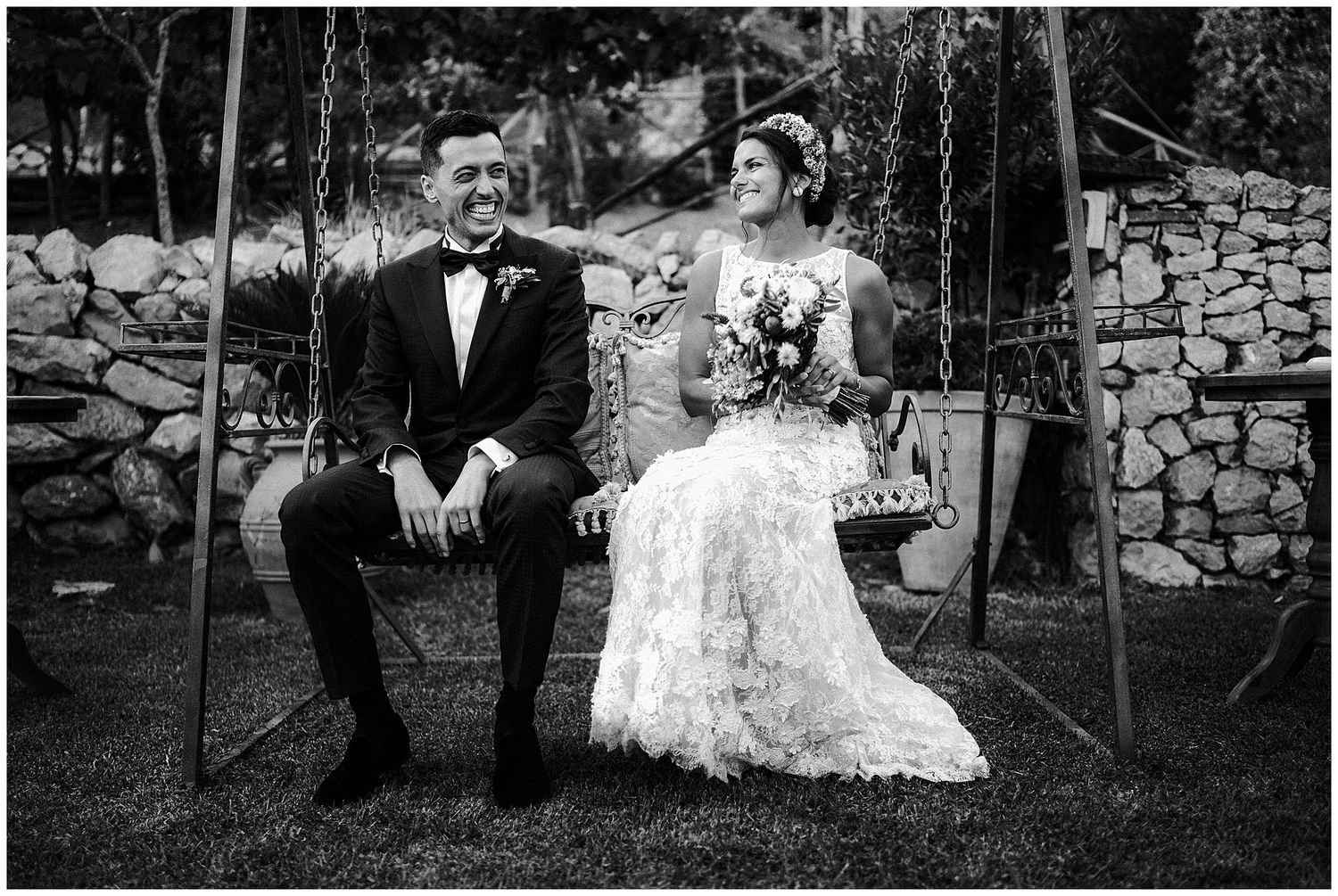 A bride and groom sitting together on a swing in front of a stone wall, captured in black and white photography.