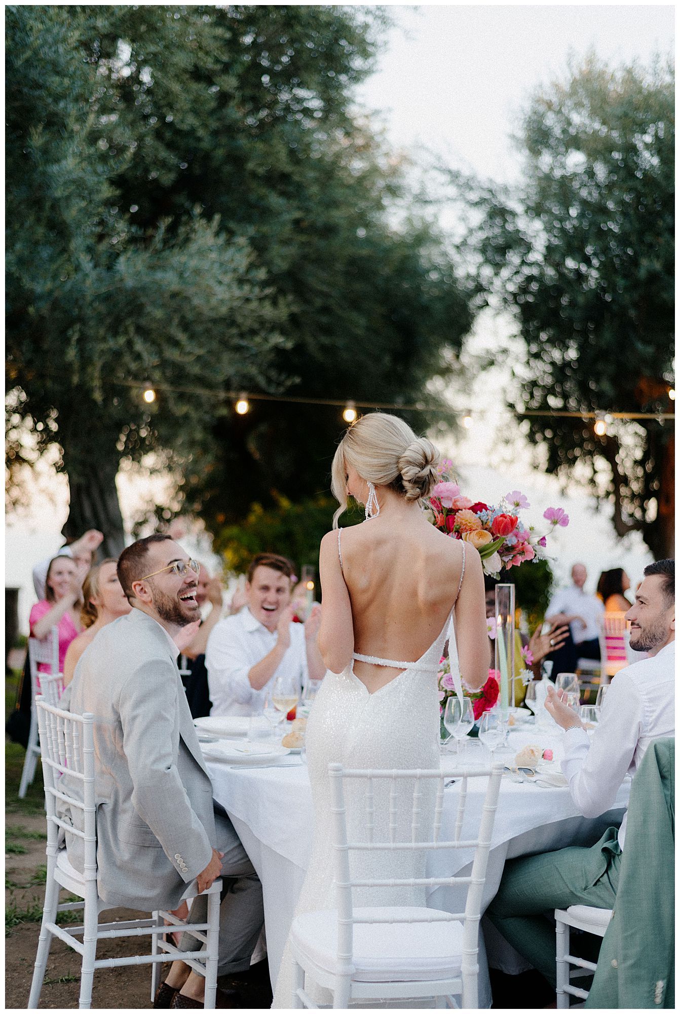 Bride in backless white dress standing at outdoor reception table while guests laugh and celebrate.