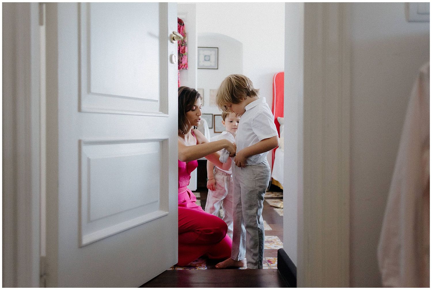 Through a doorway, two people interact in a bedroom with bright red accents, captured in soft natural light.