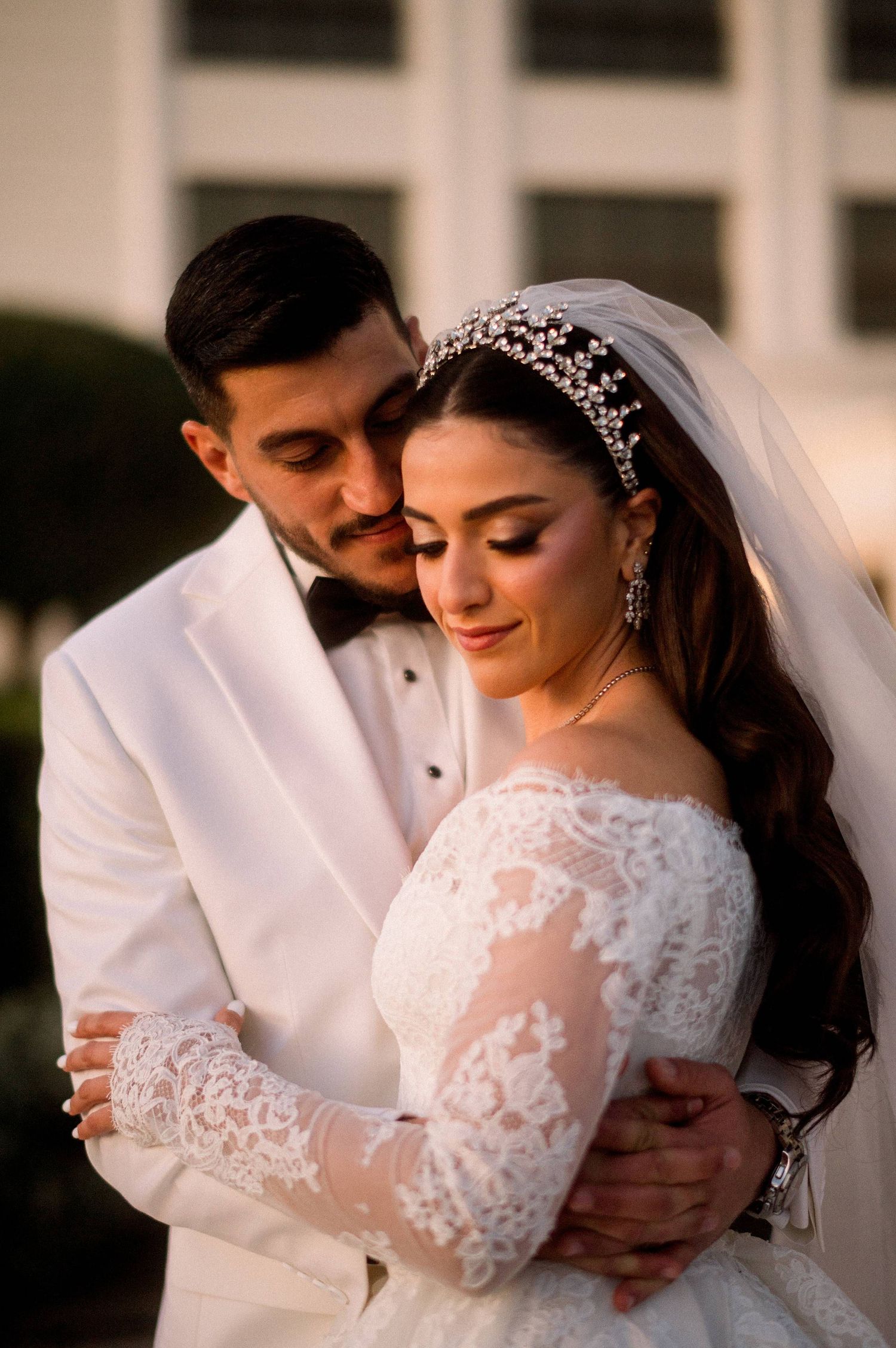 A romantic wedding portrait at sunset showing a couple embracing, with the bride in lace sleeves and crystal headpiece.