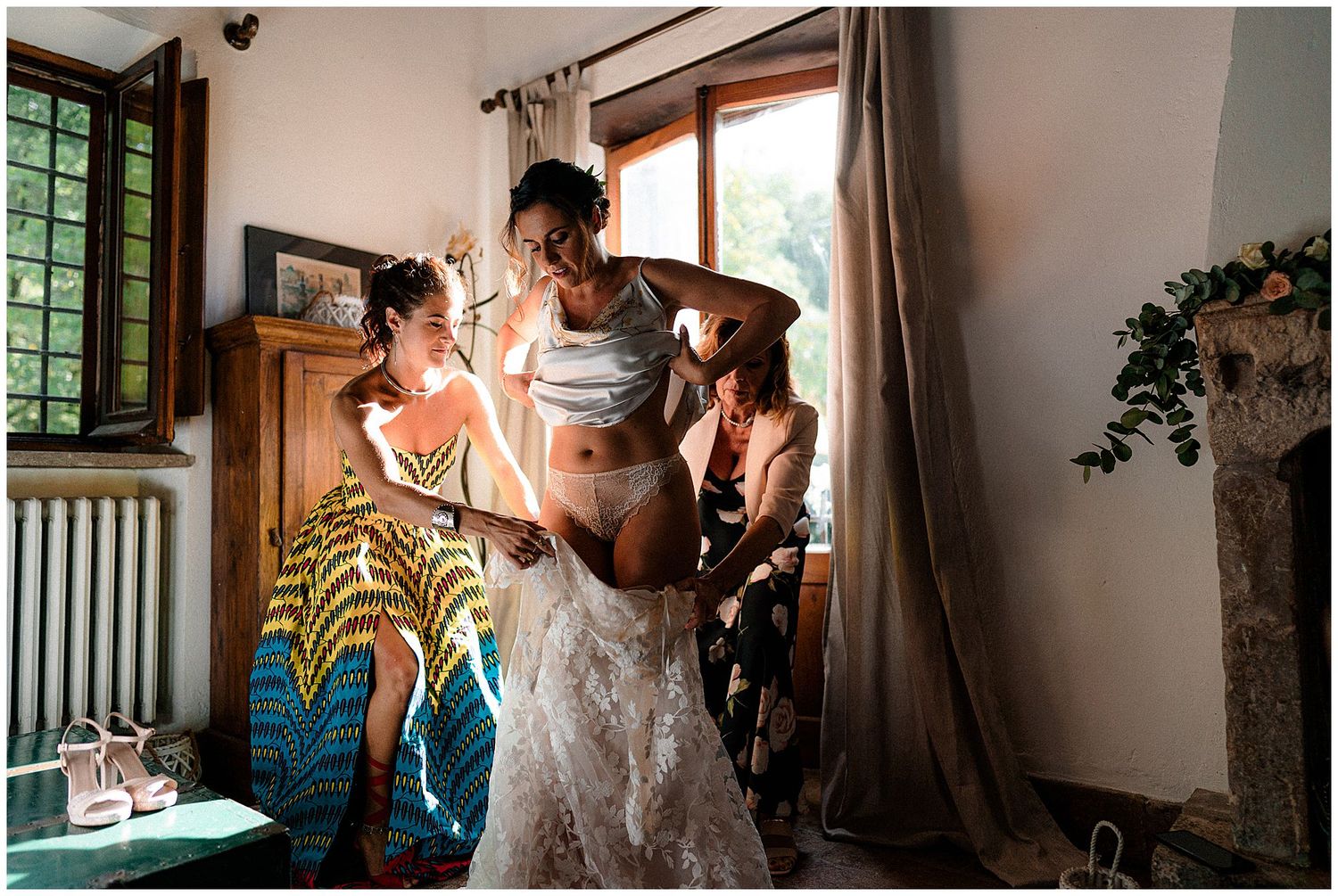 People preparing for a special event help with a white lace dress in a sunlit room with floral decor and wooden trim.