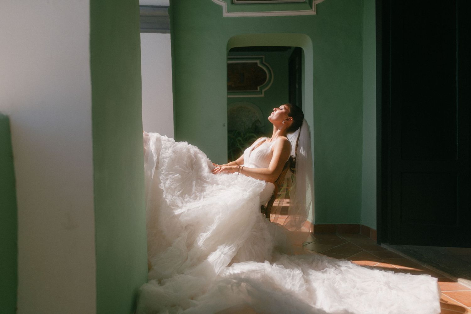 A person in a flowing white wedding gown reclines against a mint green wall in an elegant hallway with natural lighting.