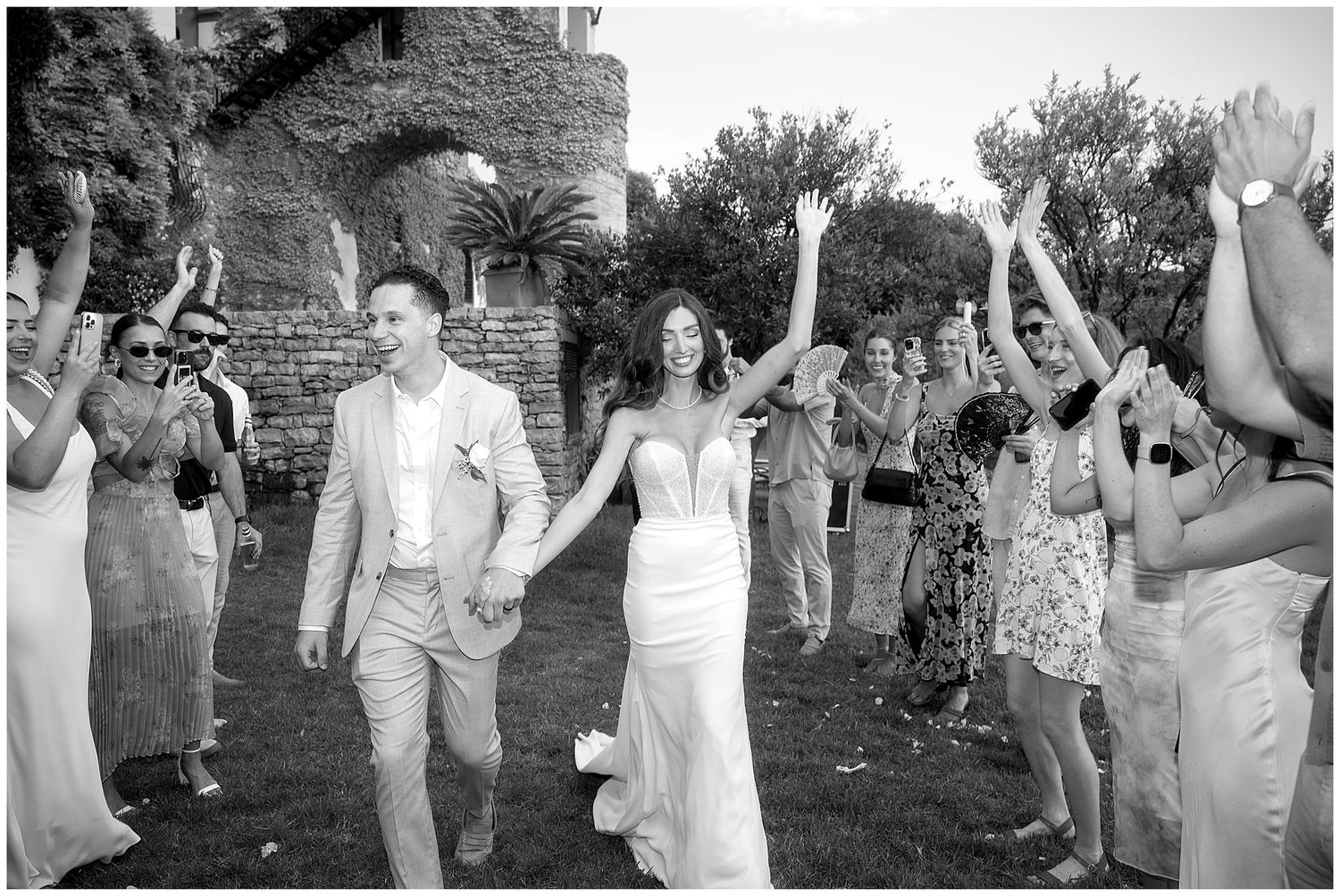 Wedding guests cheer and raise their arms in celebration as a couple exits through a joyful walkway outdoors.